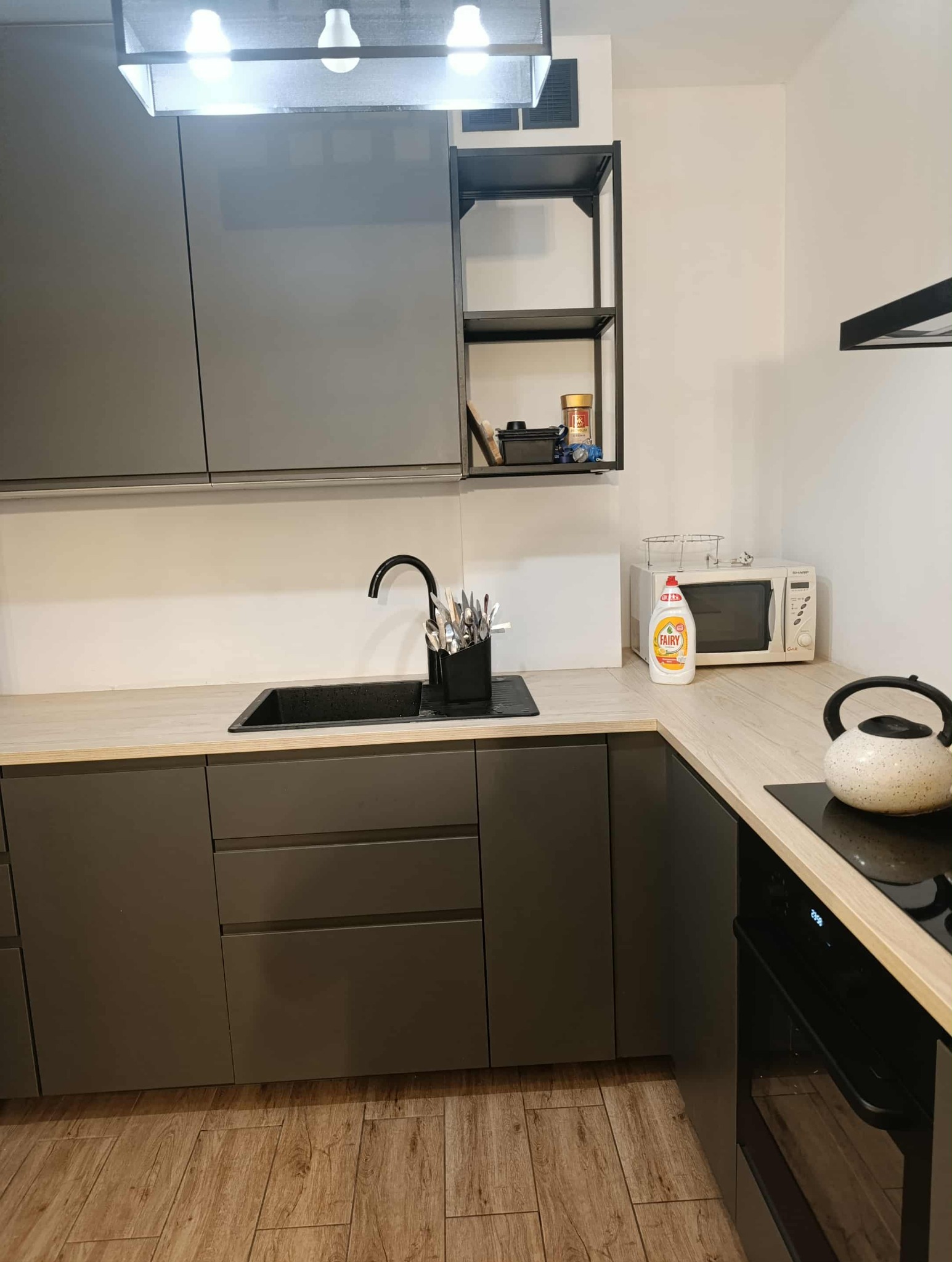 Nowoczesna, szara kuchnia z drewnianym blatem, czarnym zlewem i czajnikiem na płycie. Widoczne środki czystości i akcesoria kuchenne na półkach.