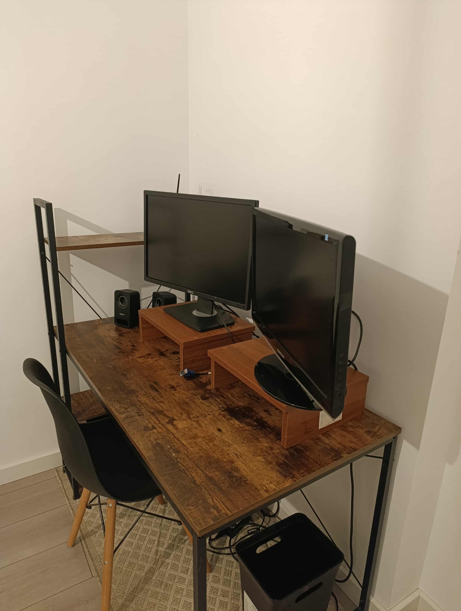 Biurko z dwoma monitorami na drewnianych podstawkach, czarne głośniki i krzesło. Widoczne kable pod biurkiem i kosz na śmieci. Minimalistyczny styl, jasne ściany.