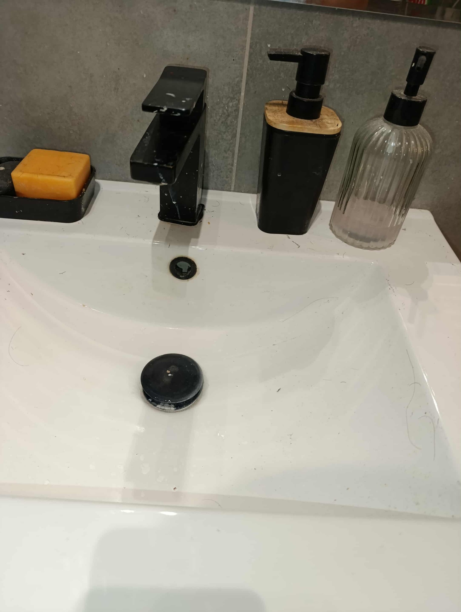 Biała umywalka z czarną armaturą, dozownikiem mydła i mydłem w kostce. Widoczne drobne zabrudzenia i włosy. Ujęcie z góry, oświetlenie naturalne.