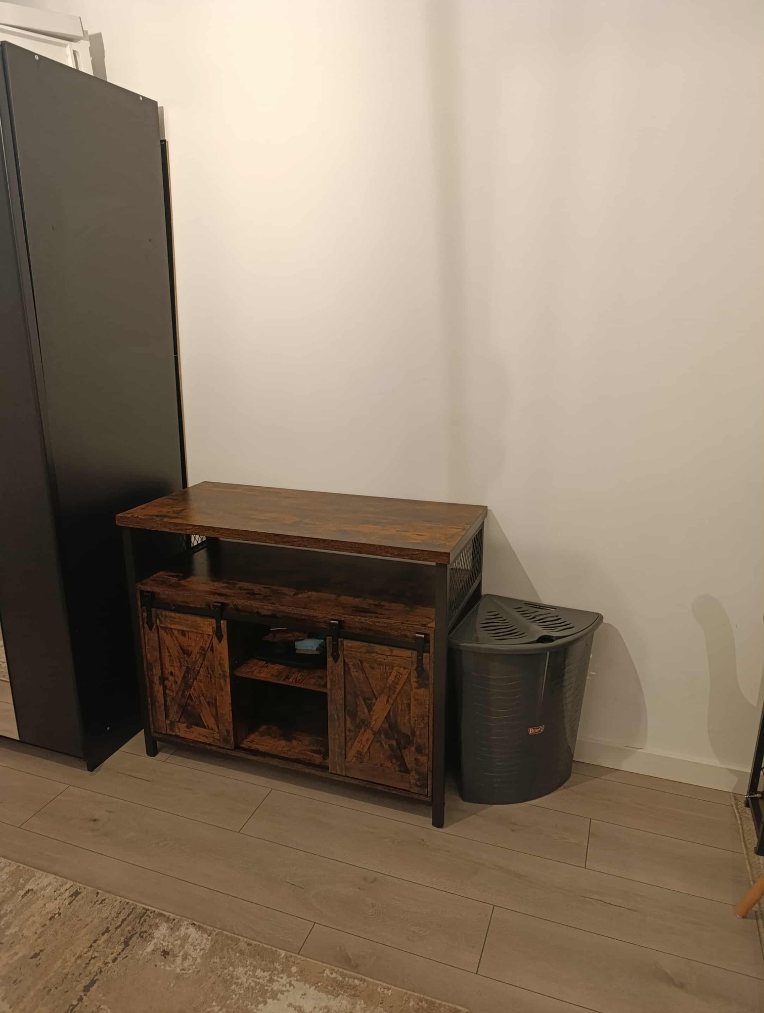 Rustykalna szafka RTV z drewnianymi drzwiczkami i metalową siatką po bokach, stojąca na podłodze obok czarnej szafy i kosza na pranie.
