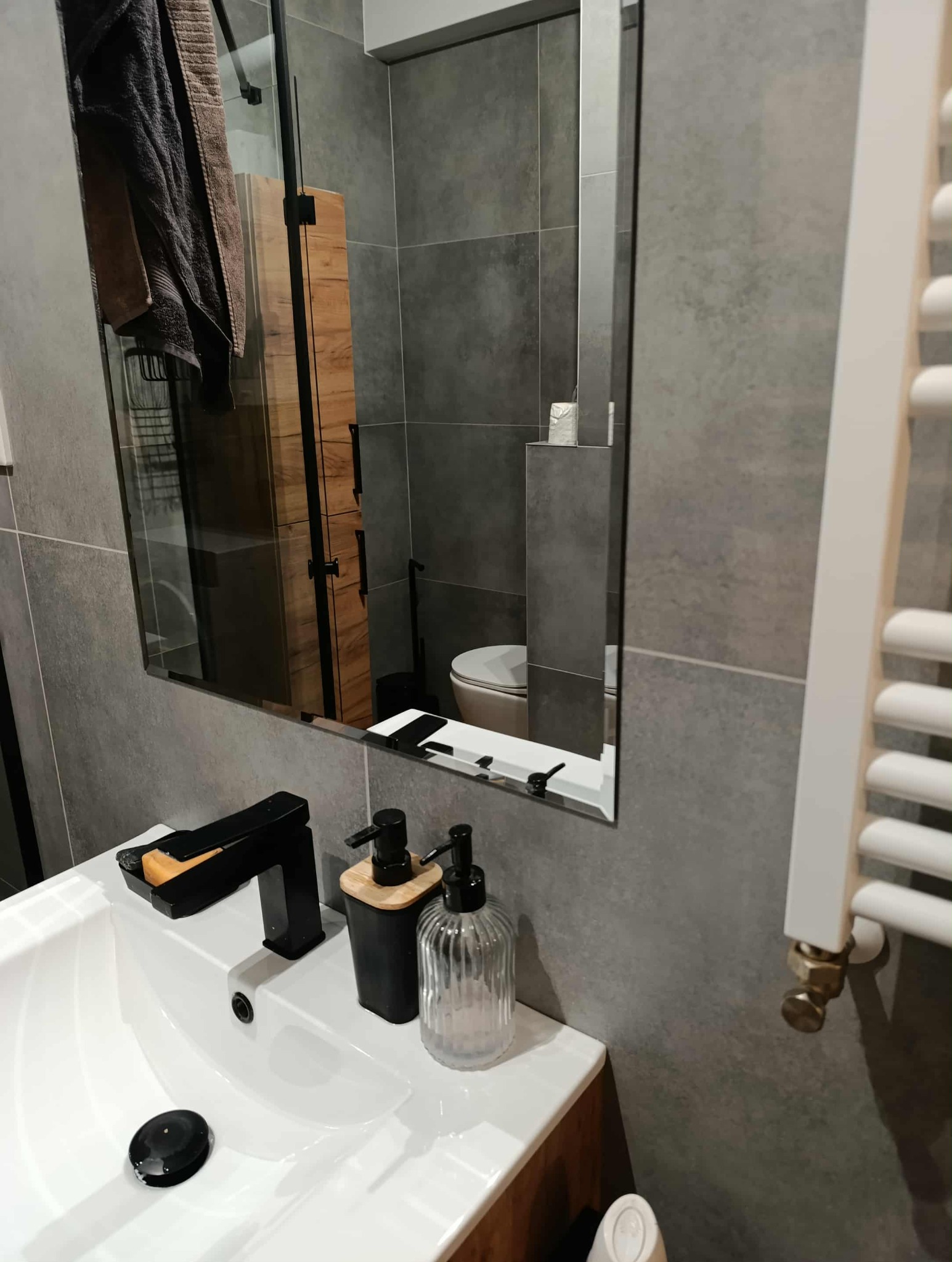 Szara łazienka po remoncie: umywalka z czarną armaturą, dozowniki mydła, lustro odbijające toaletę i grzejnik. Nowoczesne płytki, minimalistyczny design.