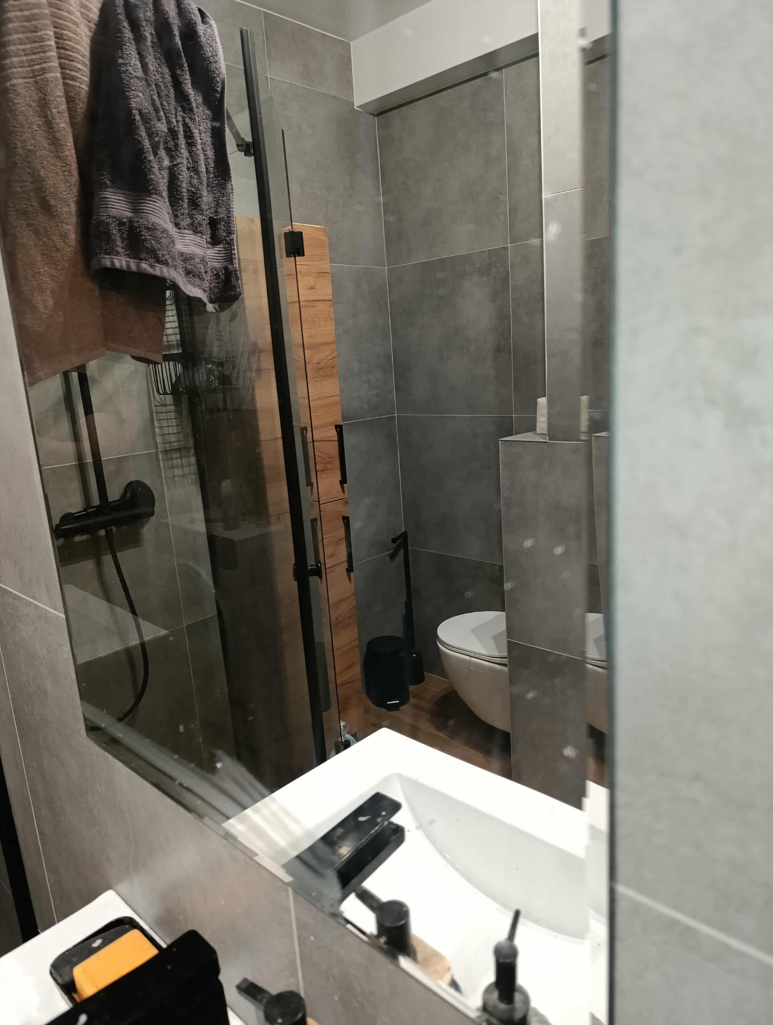 Nowoczesna łazienka z szarymi płytkami, kabiną prysznicową i białym umywalką, widziana w odbiciu lustra. Widoczne ręczniki i akcesoria łazienkowe.