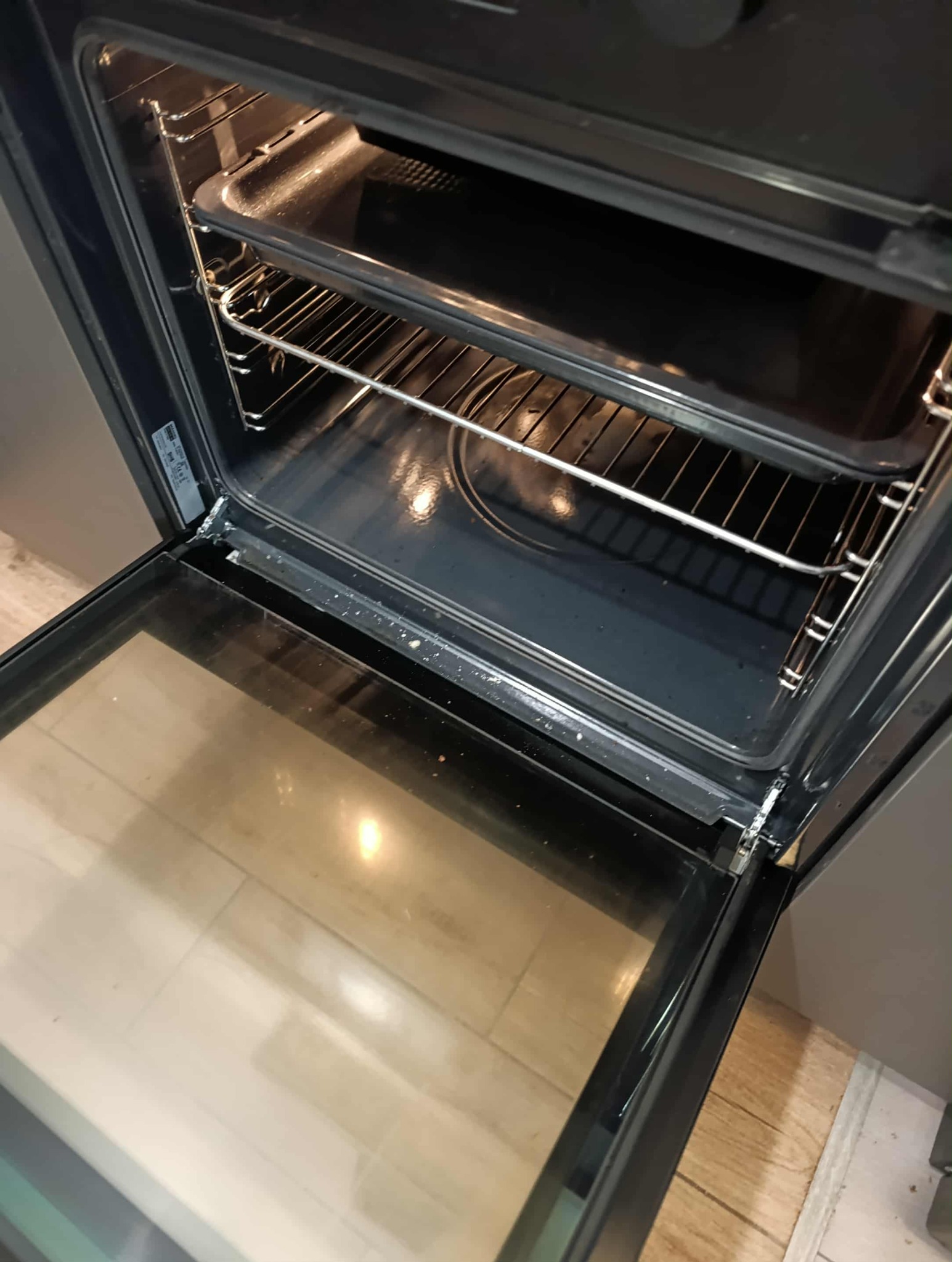 Otwarty piekarnik z widocznymi rusztami i blachą. Wnętrze piekarnika jest lekko zabrudzone, co sugeruje potrzebę czyszczenia. Widoczna szyba drzwi piekarnika.