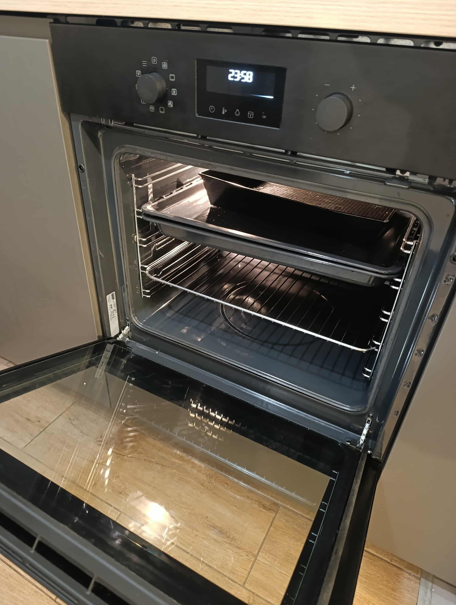 Otwarty piekarnik z rusztem i blachą w środku, widoczne wnętrze piekarnika i elementy sterujące. Ustawiony zegar na 23:58.