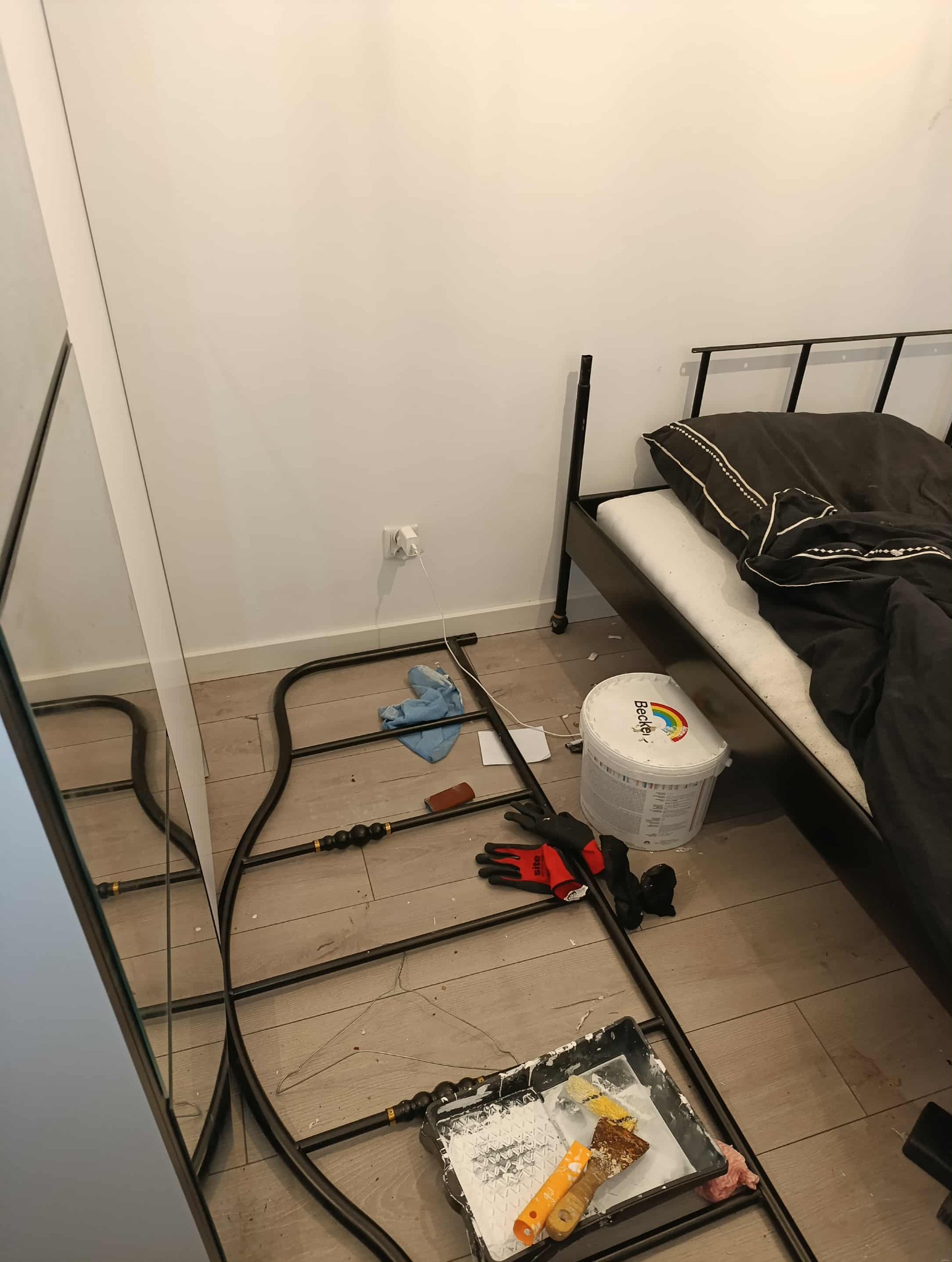 Pokój w trakcie malowania: puszka farby Becker's, kuweta z farbą, pędzle, rękawice robocze, szmatka, fragment łóżka i ramy lustra. Widoczne zabrudzenia farbą na podłodze.