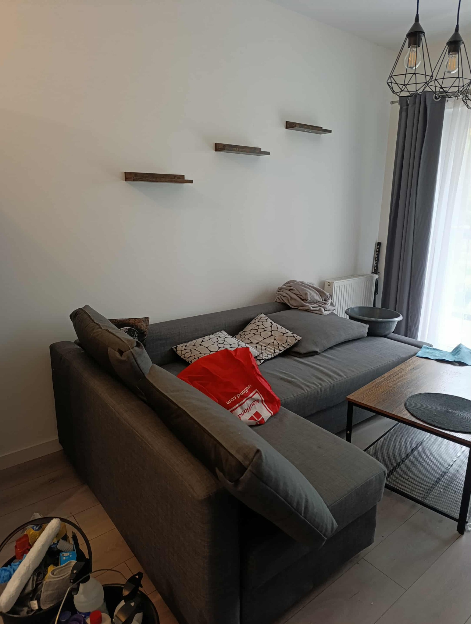 Szara narożna kanapa z poduszkami i torbą na tle białej ściany z półkami. Obok stolik kawowy i kosz ze środkami czystości. Widoczne szare zasłony.