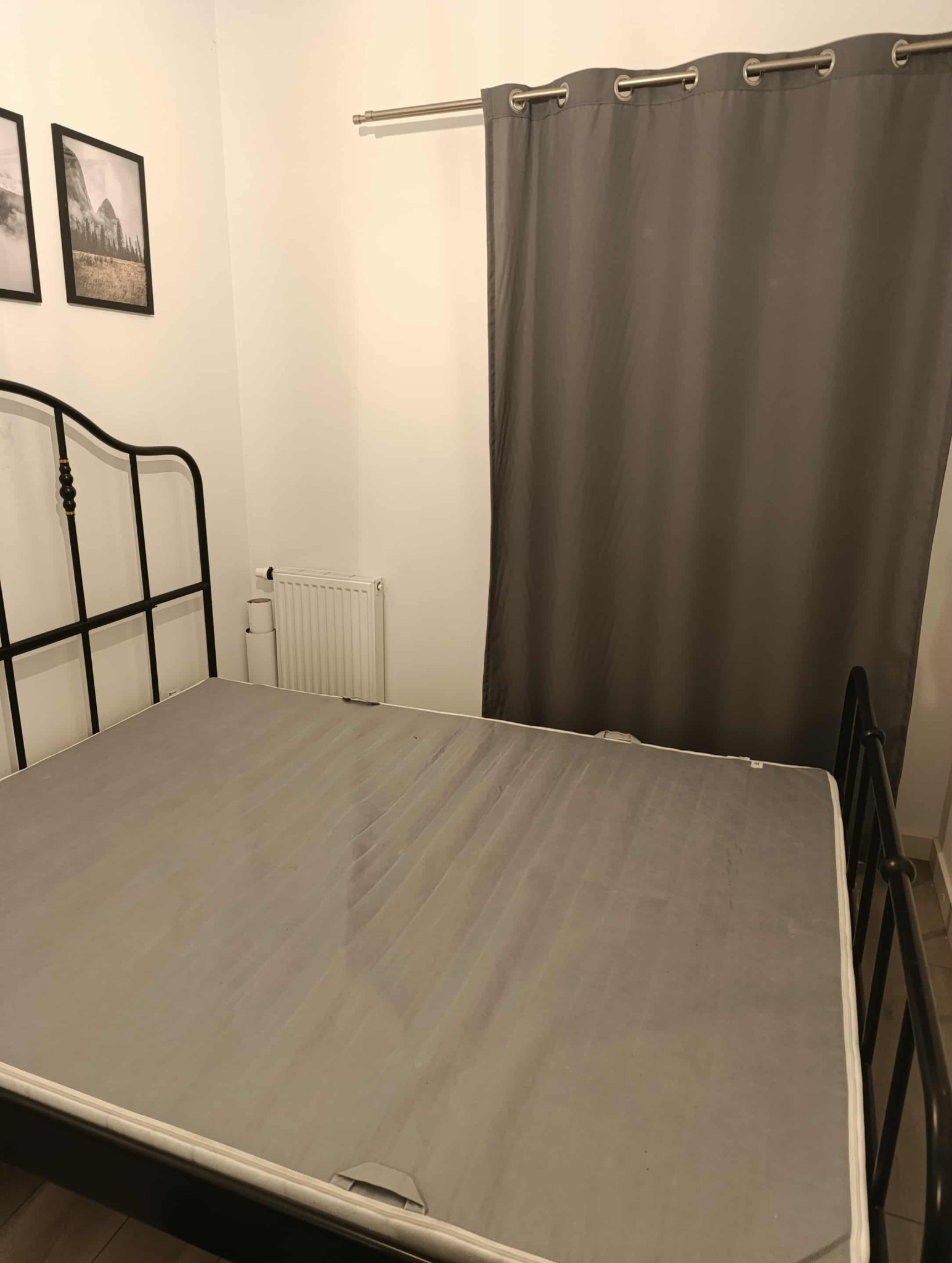 Szary materac na metalowym łóżku, widoczne odbarwienia. W tle zasłony, grzejnik i zdjęcia na ścianie.