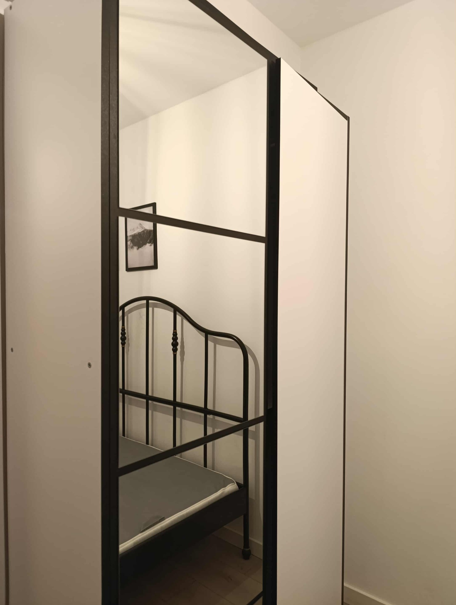 Pokój z metalowym łóżkiem widziany przez ramę szafy z lustrem. Minimalistyczny wystrój, czarno-biała kolorystyka. Na ścianie wisi obrazek w czarnej ramce.