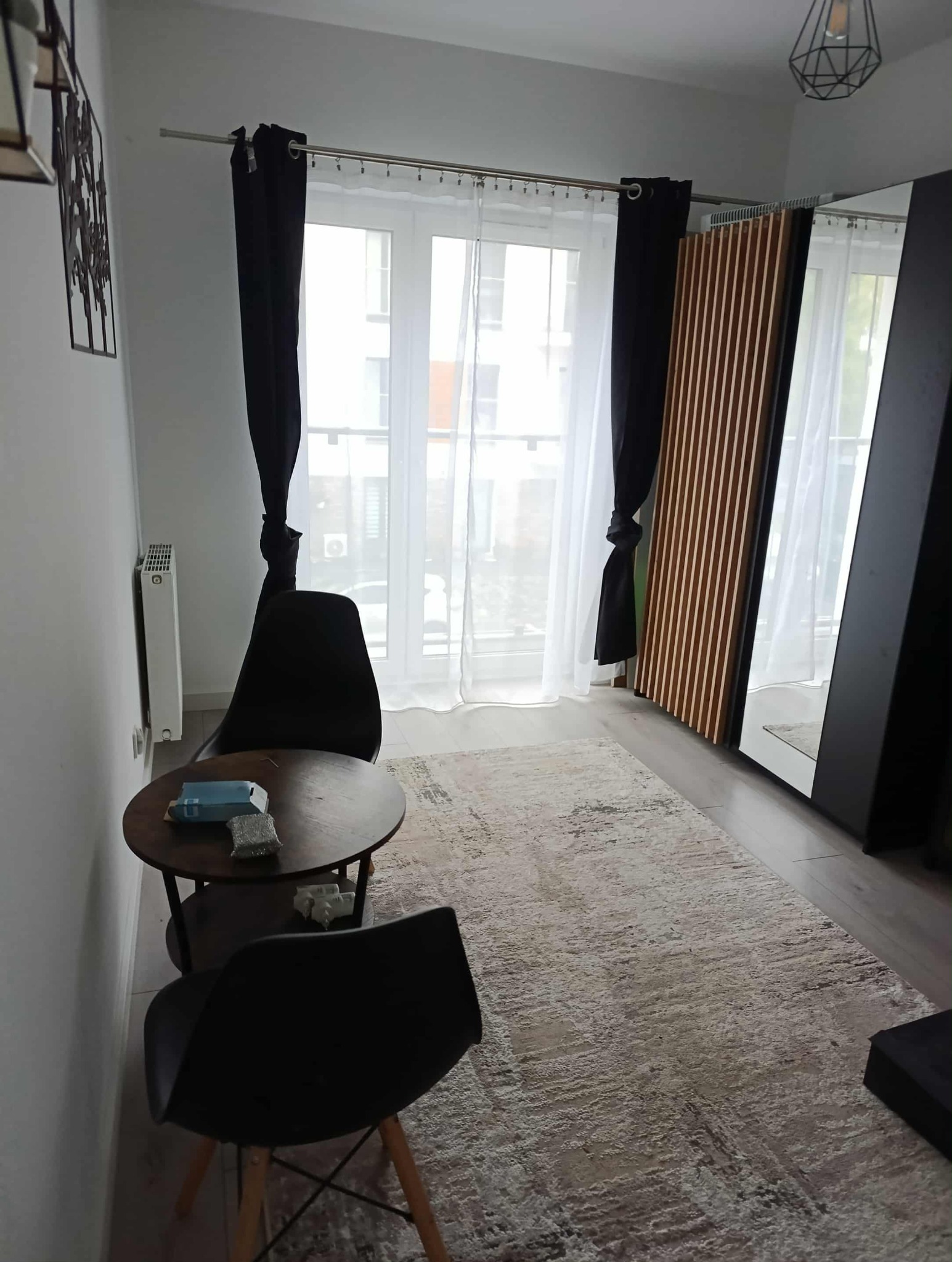 Ustawny pokój z oknem, stolikiem i dywanem w neutralnych barwach. Czarne zasłony i krzesła dodają kontrastu. Widoczne dekoracje ścienne i lampa.