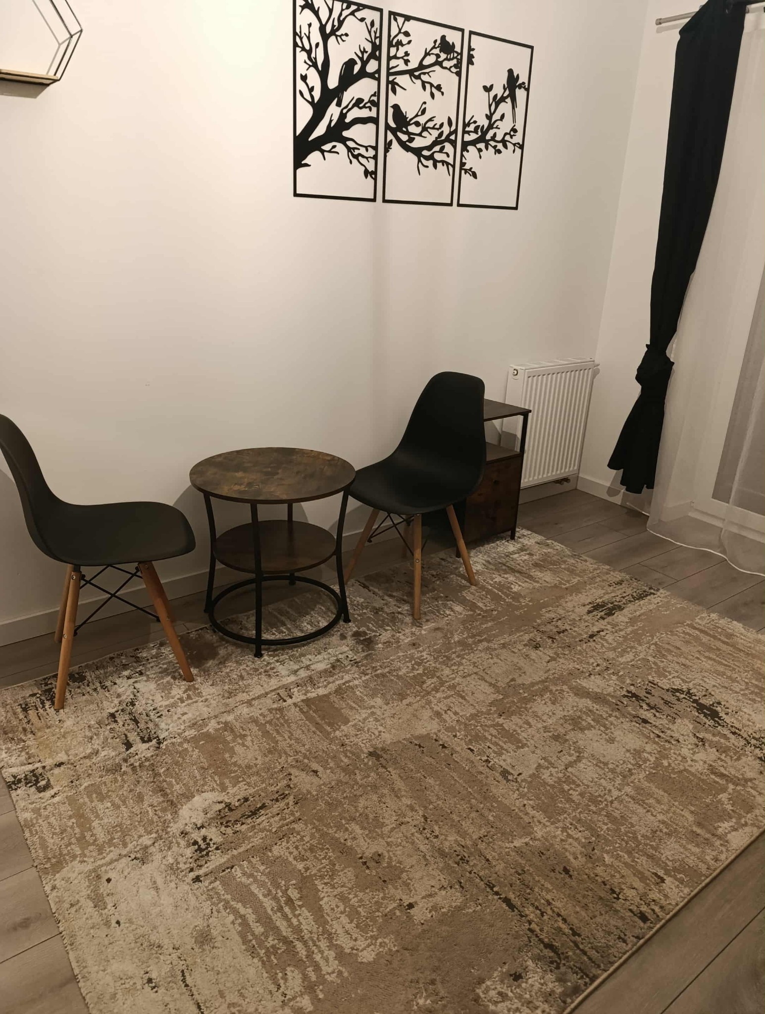 Ustawienie: Dwa krzesła i stolik na wzorzystym dywanie w pokoju z dekoracjami ściennymi. Minimalistyczny wystrój wnętrza z akcentami w kolorze czarnym i beżowym.