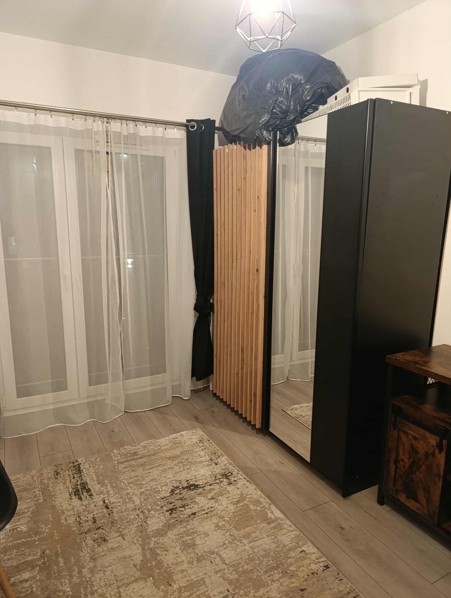 Pokój z drewnianą ścianką działową, zasłoniętymi oknami i dywanem. Na szafie czarny worek. Widoczne meble: szafa z lustrem i komoda.