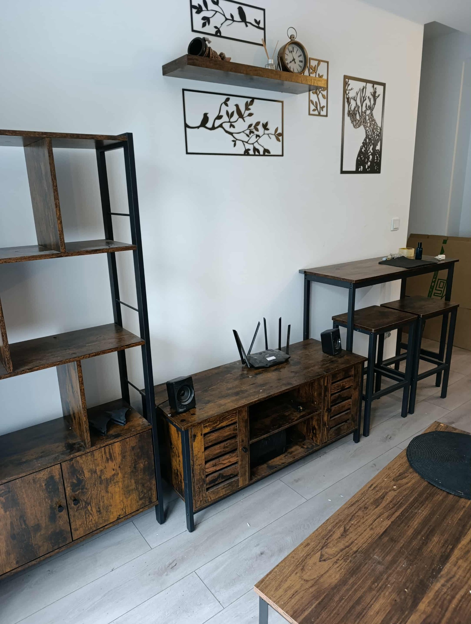 Rustykalny salon z drewnianymi meblami w stylu loft: regał, szafka RTV, stół barowy z krzesłami. Dekoracje ścienne z motywem roślinnym i zegar. Jasne panele podłogowe.