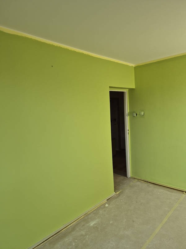 Pomalowane na zielono ściany w pokoju z widocznym wejściem do innego pomieszczenia. Podłoga zabezpieczona taśmą malarską. Widoczne niedociągnięcia przy suficie.