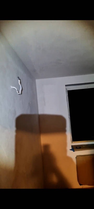 Wnętrze w trakcie remontu: szpachlowana ściana z widoczną instalacją elektryczną, okno. Ściana częściowo pomalowana na biało. Ciemne oświetlenie.