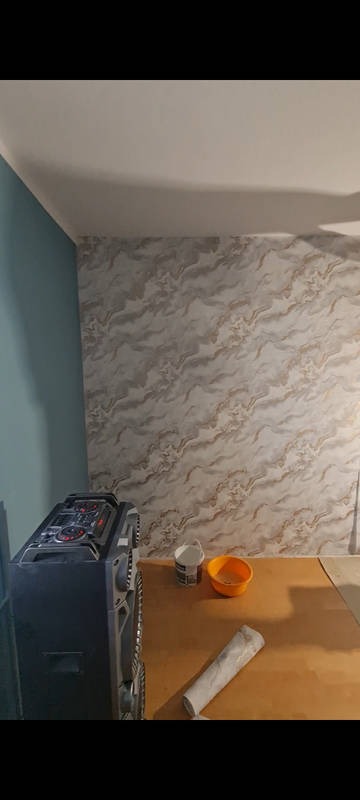 Wykończona ściana z wzorem marmuru, obok ściana pomalowana na niebiesko. Na stole wiadro farby, miska i rolka do malowania. Widoczny głośnik.