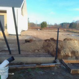 Bastion Systemy Ogrodzeniowe - Wkopane słupki i częściowo zamontowana siatka ogrodzeniowa na betonowej podmurówce. W tle widoczny dom i teren działki w trakcie prac ziemnych.