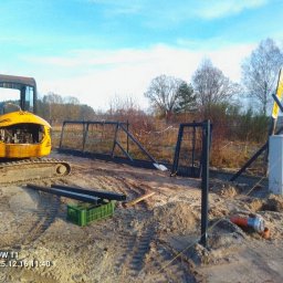 Bastion Systemy Ogrodzeniowe - Żółta mini koparka JCB obok rozłożonych elementów ogrodzenia panelowego na placu budowy, widoczne słupki i brama, w tle drzewa i błękitne niebo.