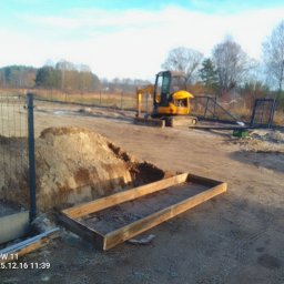 Bastion Systemy Ogrodzeniowe - Przygotowanie terenu pod ogrodzenie: wykop, drewniana rama, siatka ogrodzeniowa w tle, mała koparka. Etap prac ziemnych i fundamentowych pod budowę ogrodzenia.