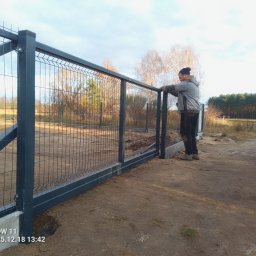 Bastion Systemy Ogrodzeniowe - Montaż panelowego ogrodzenia posesji z podmurówką. Mężczyzna w roboczym ubraniu opiera się o górną część ogrodzenia. Widok na las w tle.