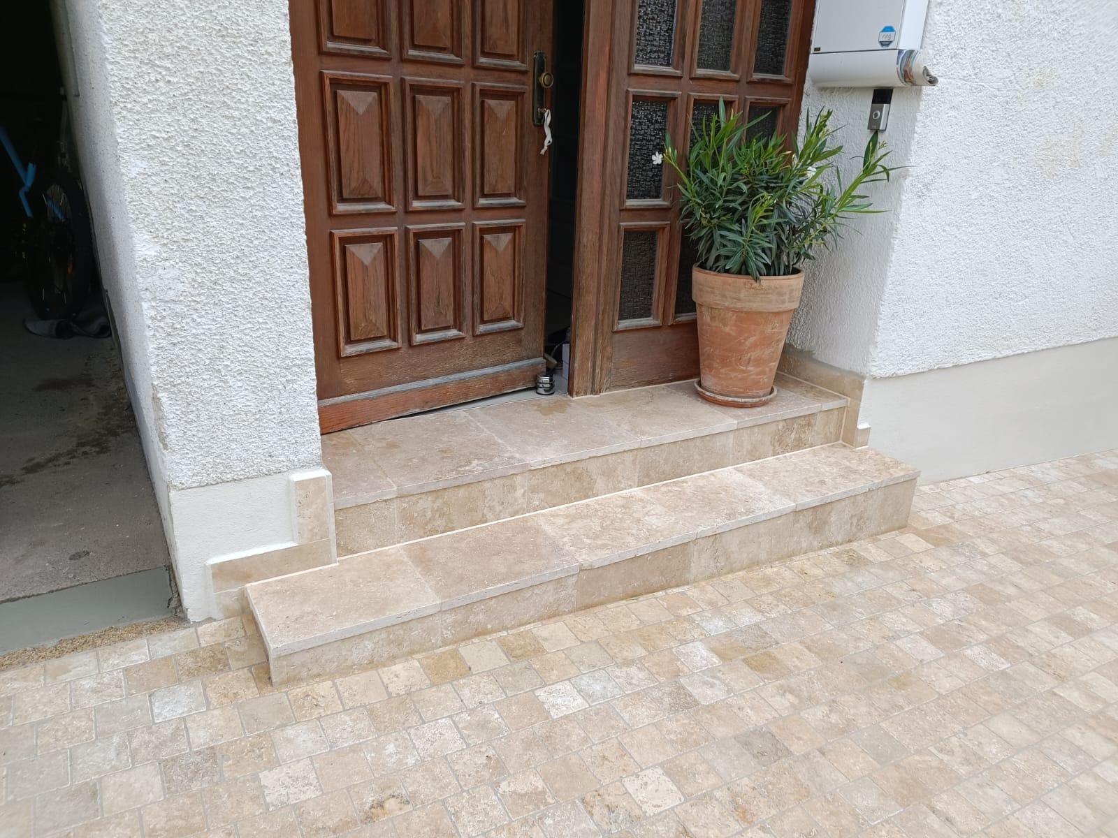 Wykończone płytkami schody wejściowe z jasnego trawertynu, prowadzące do drewnianych drzwi. Po prawej donica z zieloną rośliną, całość w neutralnej kolorystyce.