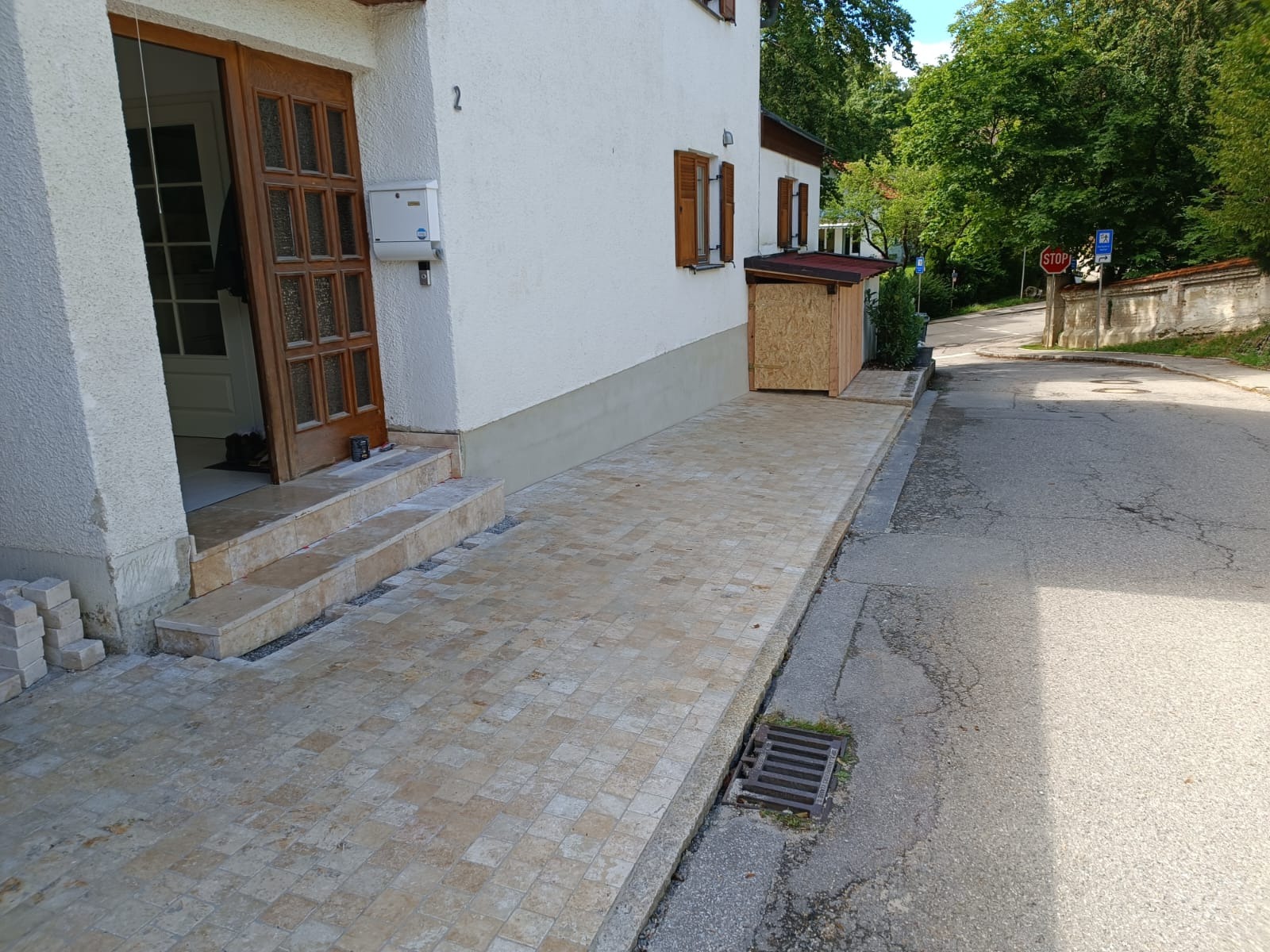 Ułożona kostka brukowa tworząca chodnik i schody wejściowe do domu z numerem 2. Widoczny fragment ulicy, drzewa i znak STOP.