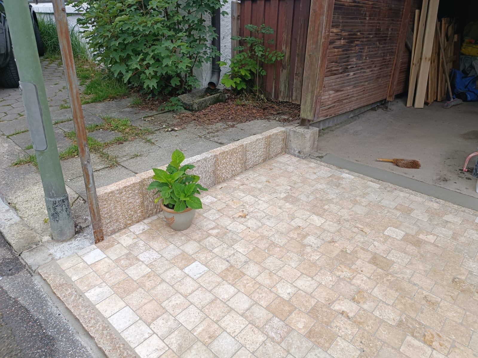 Wykończony podjazd z kostki brukowej w odcieniach beżu, otoczony niskim murkiem z kamienia. W tle widoczny budynek gospodarczy i zieleń.