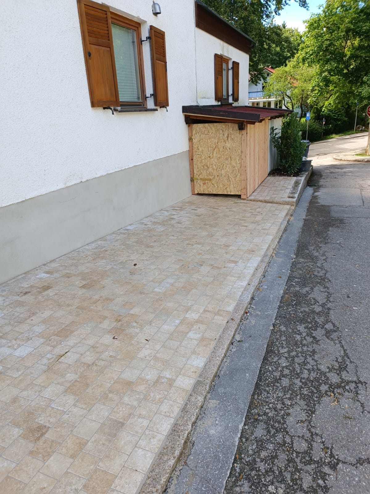 Drewniana konstrukcja przy budynku z brązowymi okiennicami, wyłożona beżową kostką brukową. Elementy z płyty OSB.