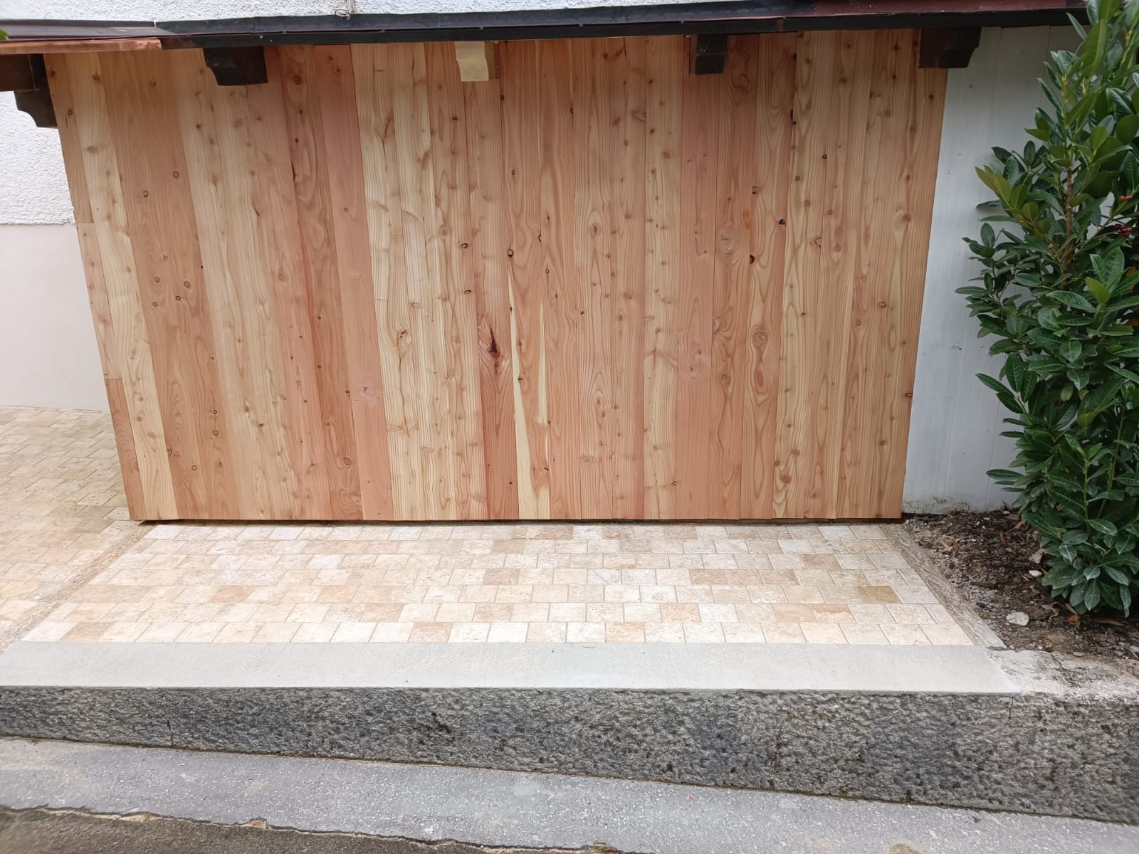 Drewniana osłona śmietnika na zewnątrz, wykonana z pionowo ułożonych desek, z widocznym fragmentem dachu i roślinnością obok, na tle jasnej elewacji.