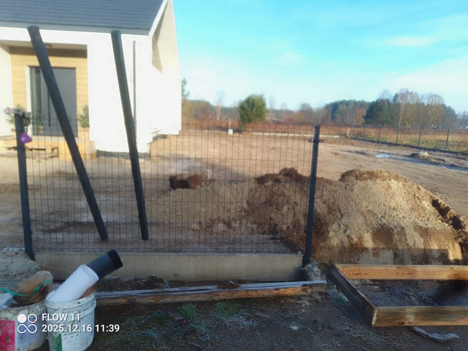 Wkopane słupki i częściowo zamontowana siatka ogrodzeniowa na betonowej podmurówce. W tle widoczny dom i teren działki w trakcie prac ziemnych.