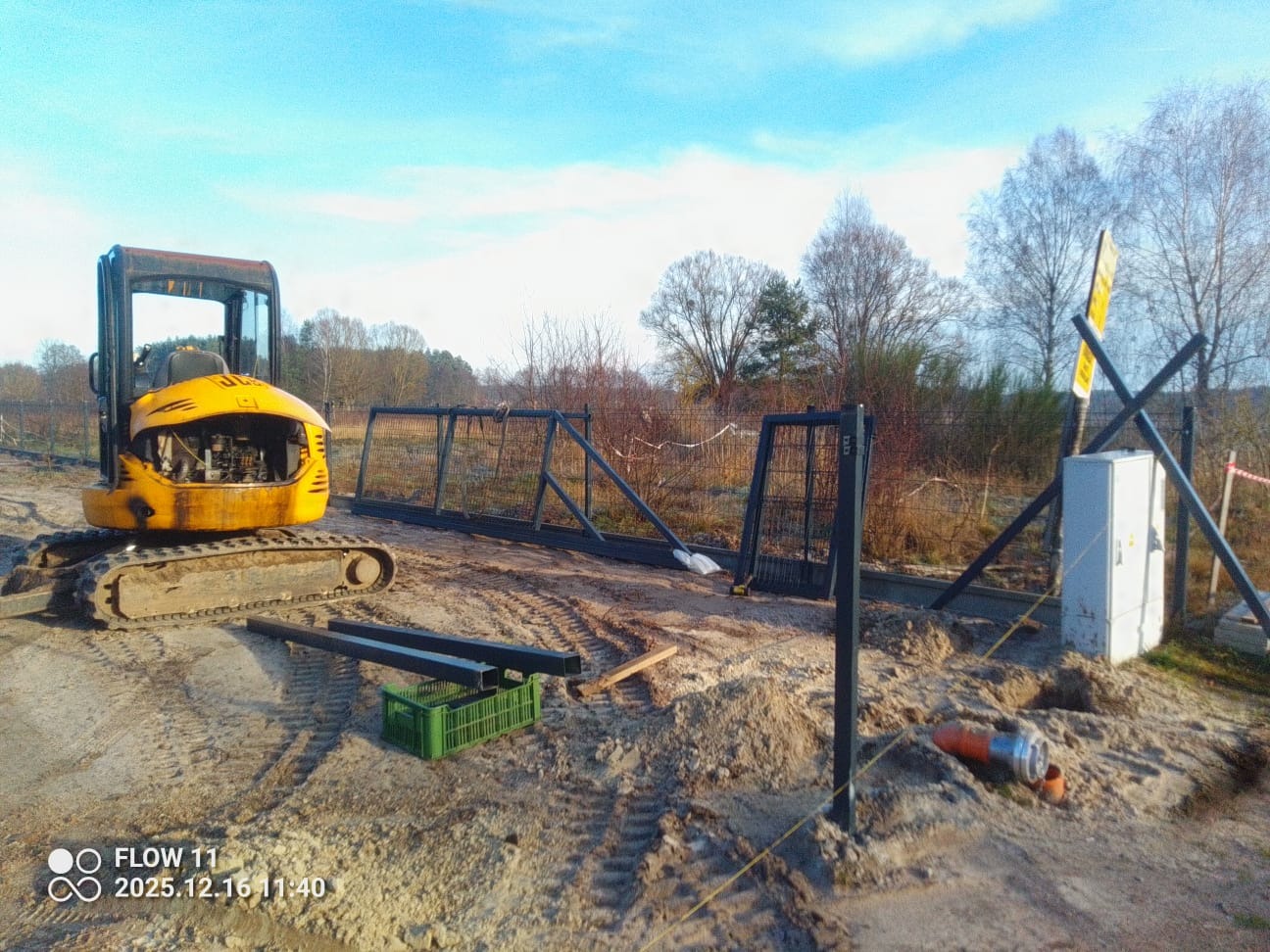 Żółta mini koparka JCB obok rozłożonych elementów ogrodzenia panelowego na placu budowy, widoczne słupki i brama, w tle drzewa i błękitne niebo.