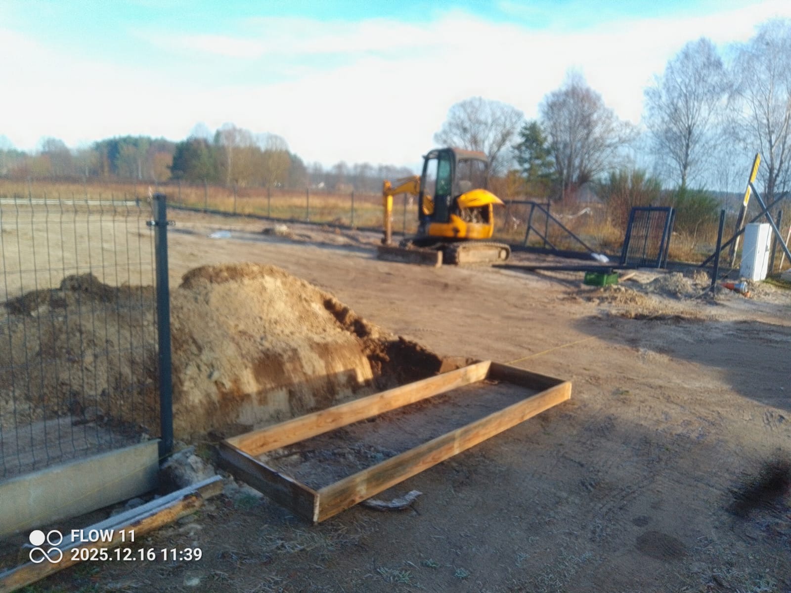 Przygotowanie terenu pod ogrodzenie: wykop, drewniana rama, siatka ogrodzeniowa w tle, mała koparka. Etap prac ziemnych i fundamentowych pod budowę ogrodzenia.