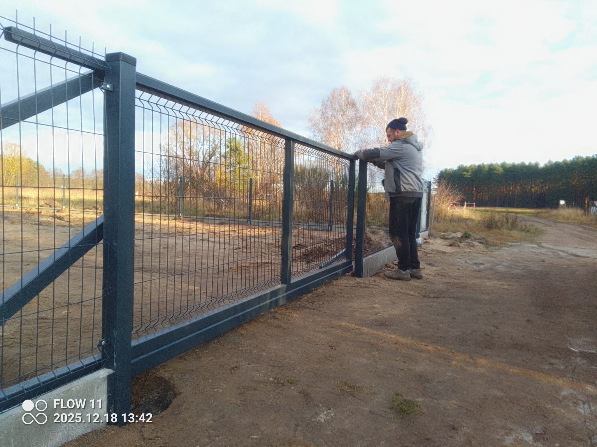 Montaż panelowego ogrodzenia posesji z podmurówką. Mężczyzna w roboczym ubraniu opiera się o górną część ogrodzenia. Widok na las w tle.