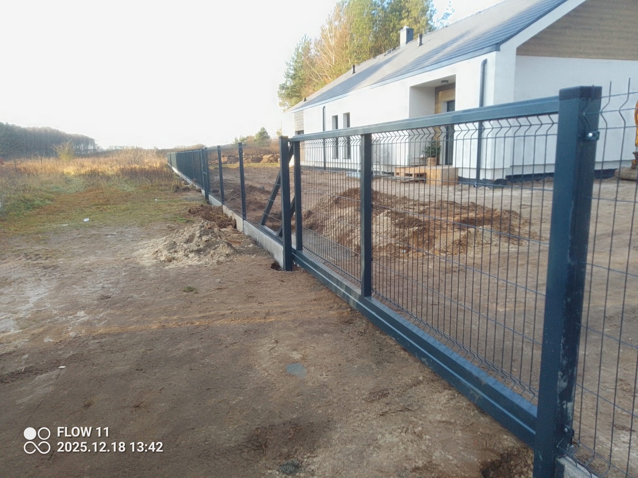 Nowoczesne, grafitowe ogrodzenie panelowe z podmurówką na tle białego domu z ciemnym dachem. Widoczne słupki i siatka ogrodzeniowa. Teren wokół ogrodzenia przygotowany pod zasiew.