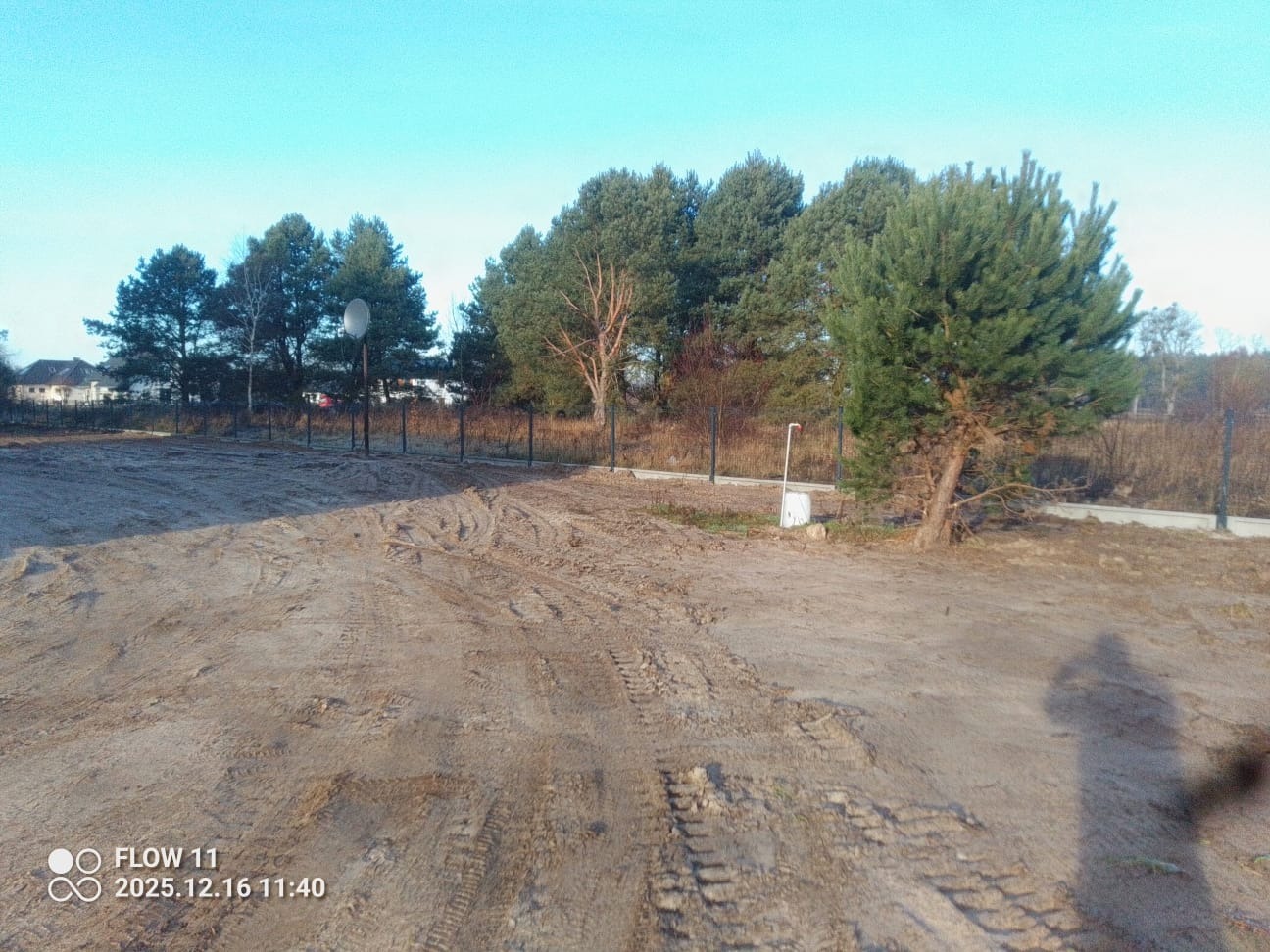 Nowoczesne ogrodzenie panelowe z metalowymi słupkami na nierównym terenie, w tle drzewa i budynek. Widoczne ślady opon na ziemi.