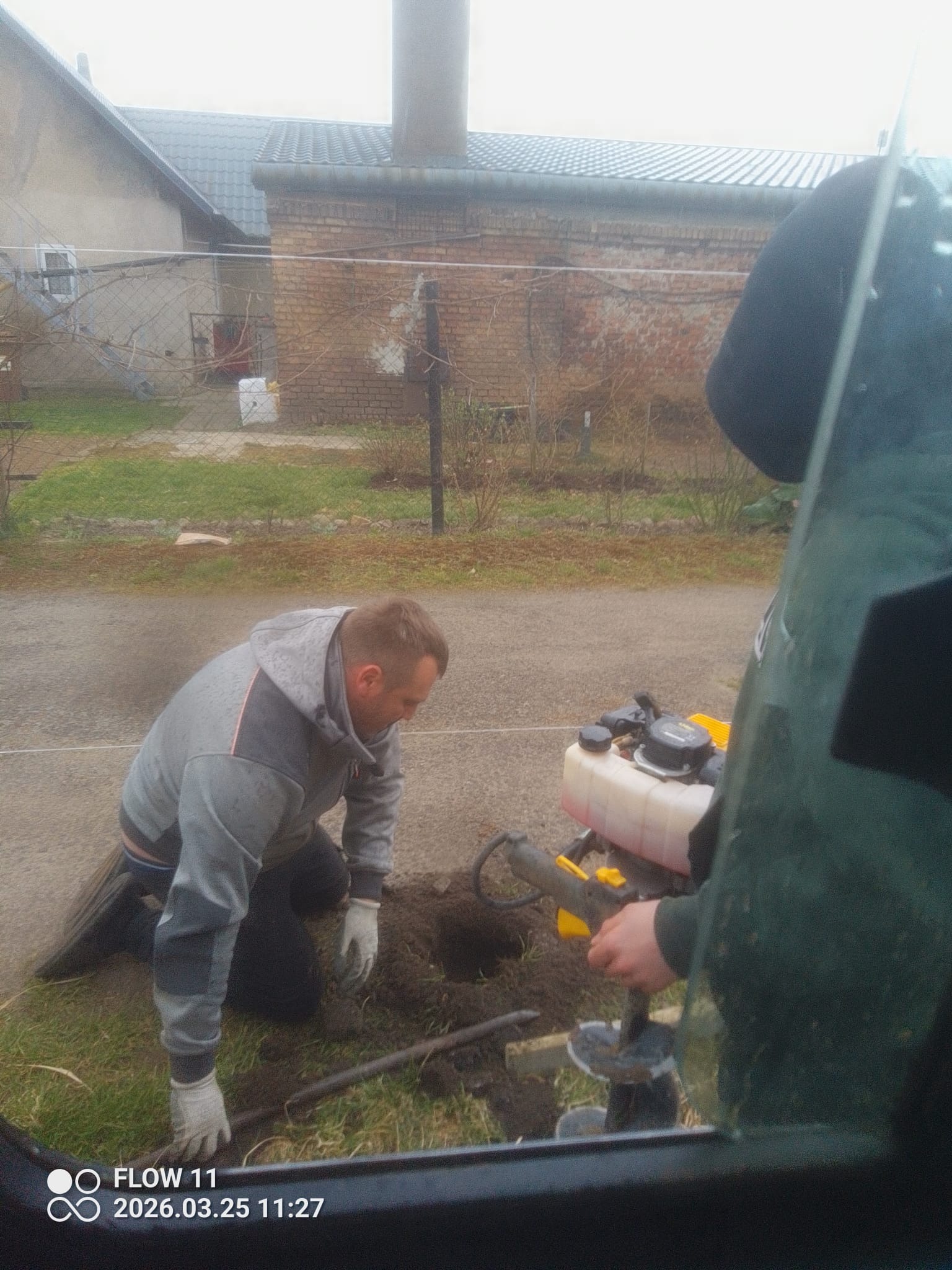 Pracownik w trakcie wiercenia otworów w ziemi w celu montażu słupków ogrodzeniowych. Widoczna wiertnica spalinowa i częściowo ogrodzenie w tle.