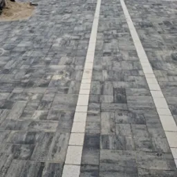 Szara kostka brukowa ułożona wzdłuż ścieżki z białymi pasami, widok z góry, lekka perspektywa, w tle zieleń drzew i zaparkowany samochód.