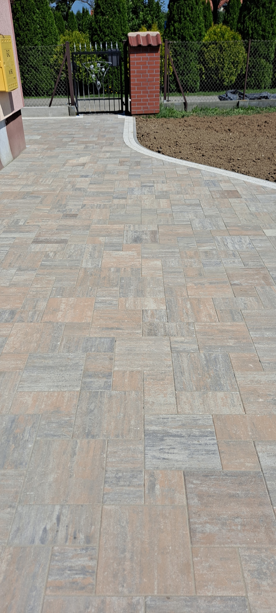 Ułożona kostka brukowa w odcieniach szarości i beżu tworzy wzór na podjeździe. W tle brama z metalu i mur z cegły. Z prawej strony widoczny przygotowany teren pod nasadzenia.