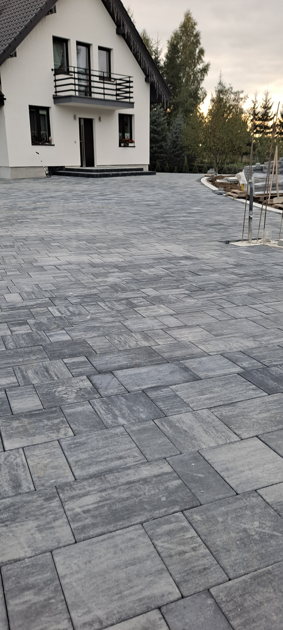 Ułożona szara kostka brukowa na podjeździe przed domem z balkonem. Fragment kostki przygotowany do dalszego układania. Widoczne narzędzia i materiały budowlane.