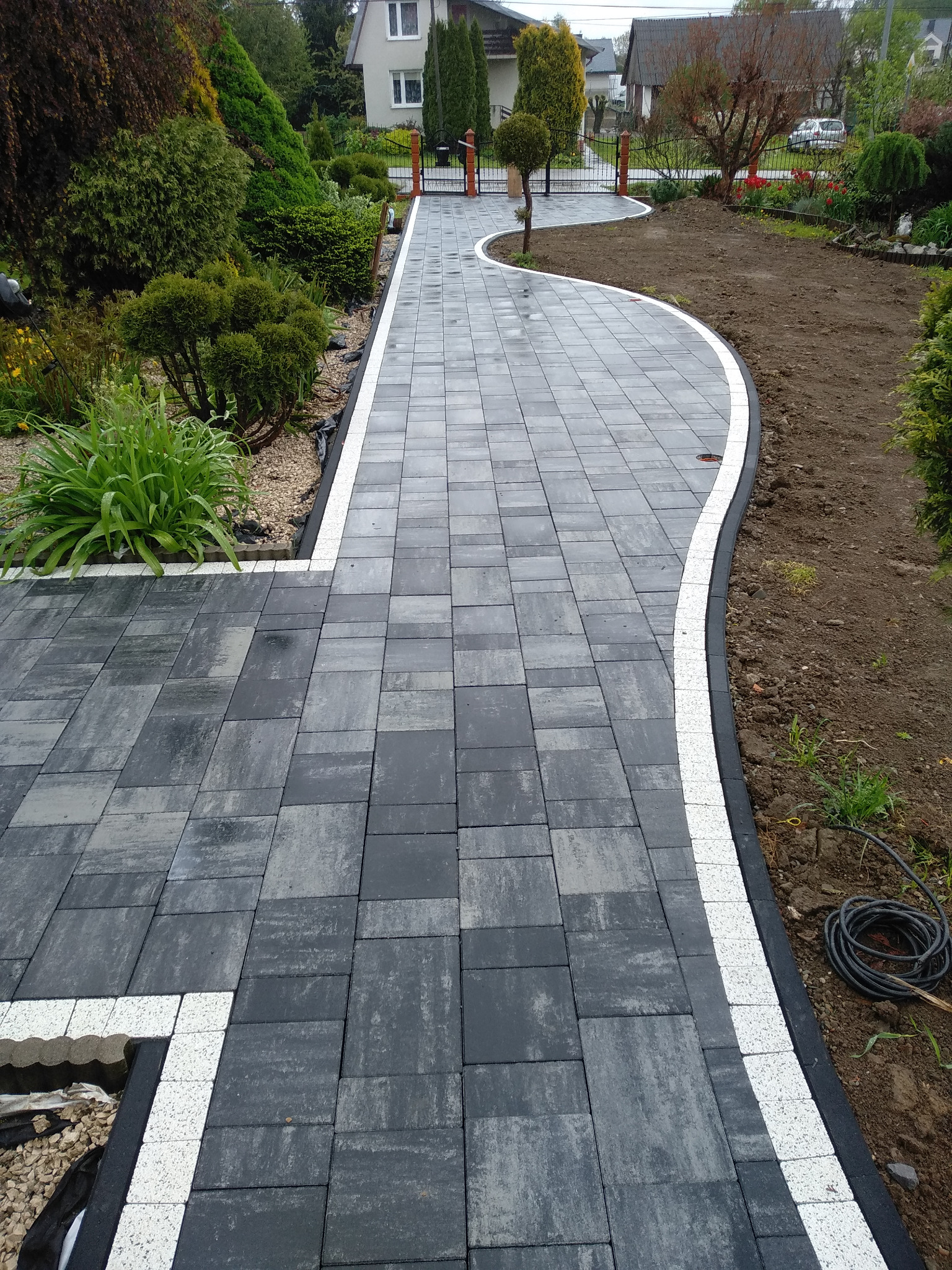 Ścieżka z szarej kostki brukowej z białym obramowaniem, wijąca się przez ogród. Widoczne świeżo przygotowane rabaty i zadbana roślinność.