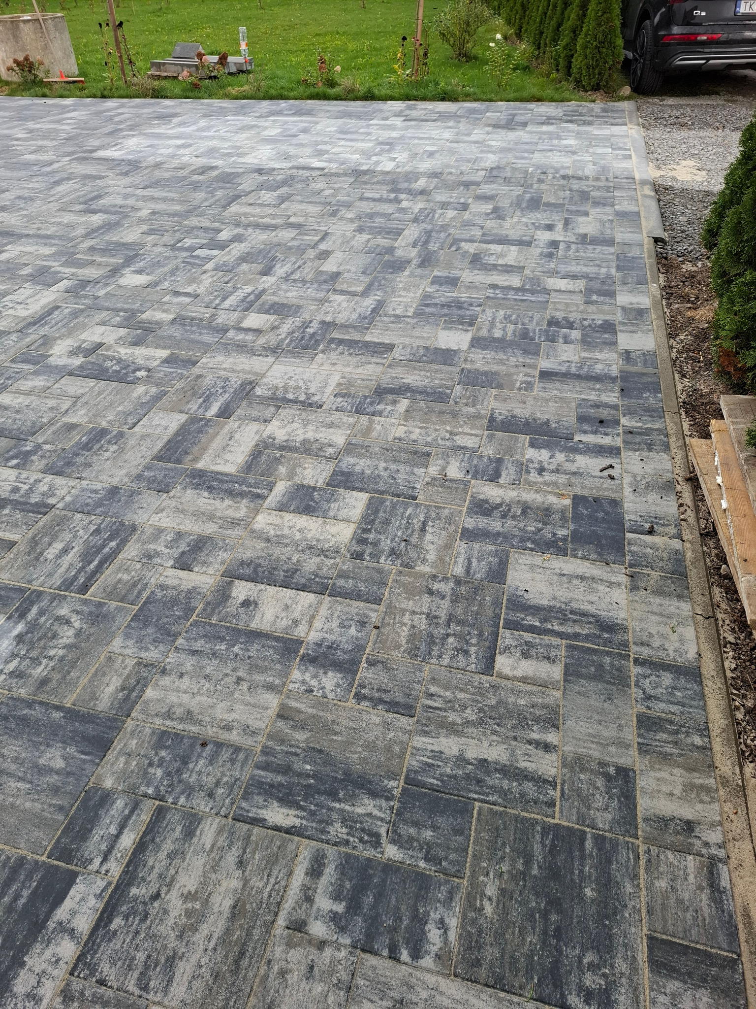 Ułożona kostka brukowa w odcieniach szarości, tworząca wzór na podjeździe. Widoczne trawnik i krzewy w tle, oraz fragment samochodu osobowego.