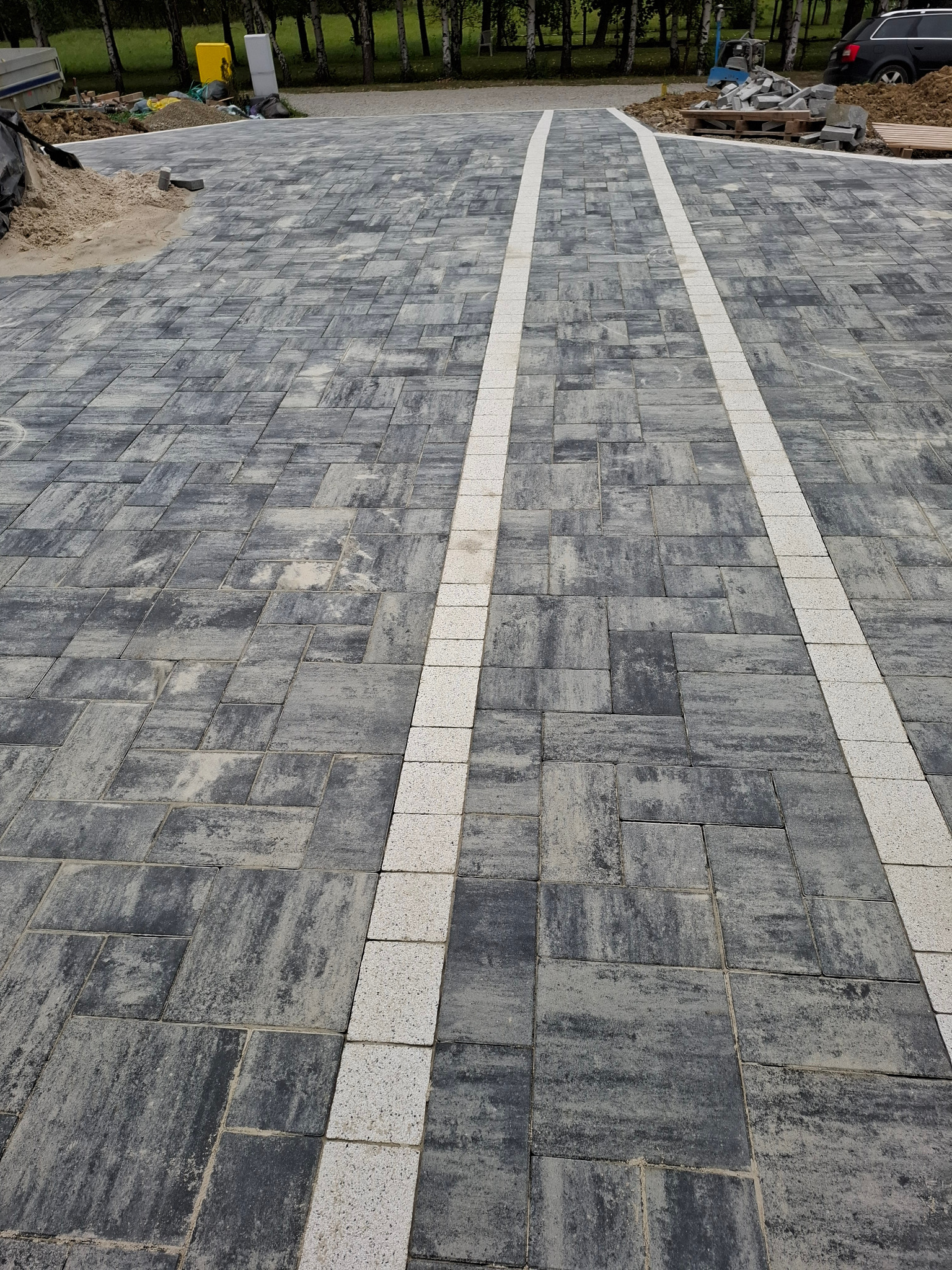 Szara kostka brukowa ułożona wzdłuż ścieżki z białymi pasami, widok z góry, lekka perspektywa, w tle zieleń drzew i zaparkowany samochód.