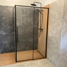 Nowoczesna kabina prysznicowa z czarnymi profilami, drewnianą podłogą i szarymi płytkami imitującymi lastryko na ścianach. Minimalistyczny design.