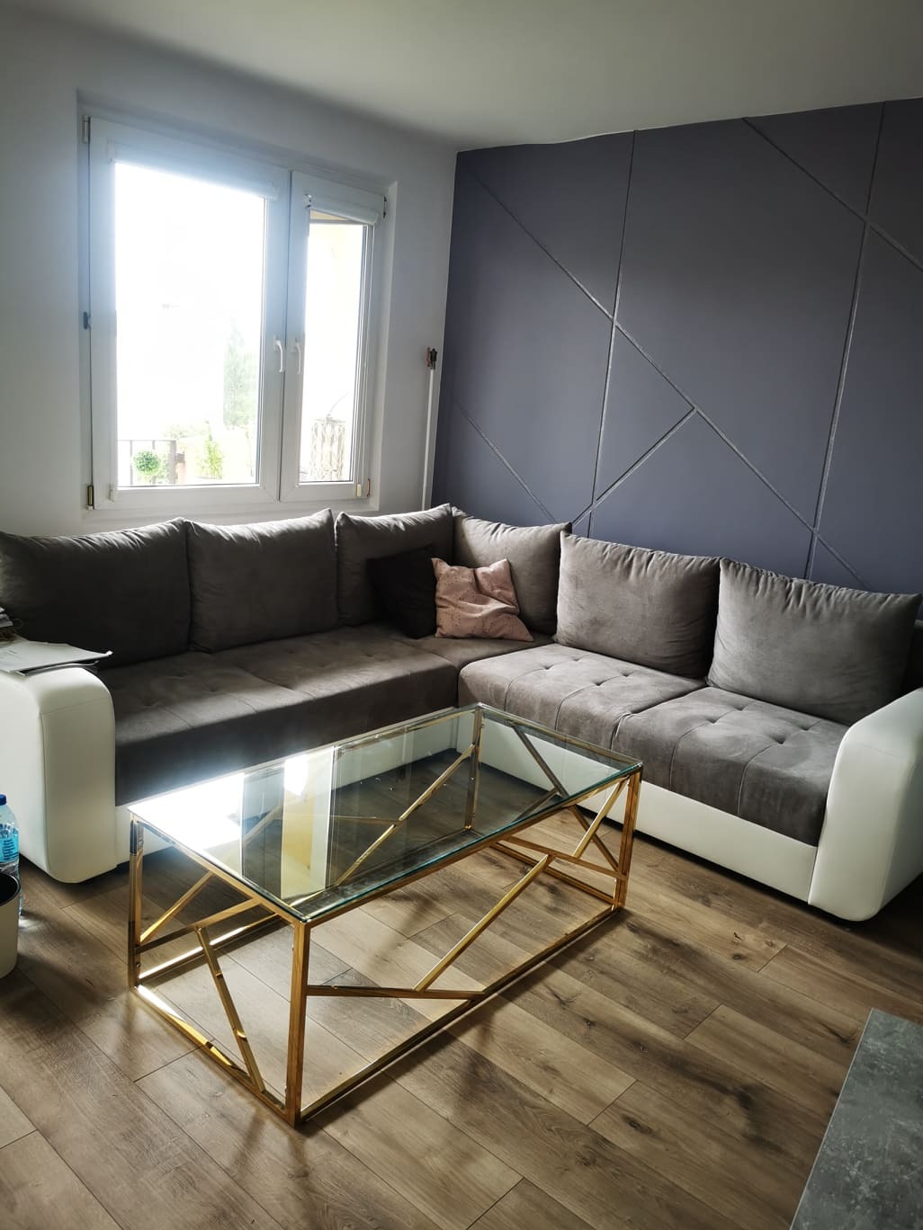 Stylowy salon z szarą ścianą w geometryczne wzory, brązowym narożnikiem i złotym stolikiem kawowym na drewnianej podłodze. Jasne, naturalne światło z okna.