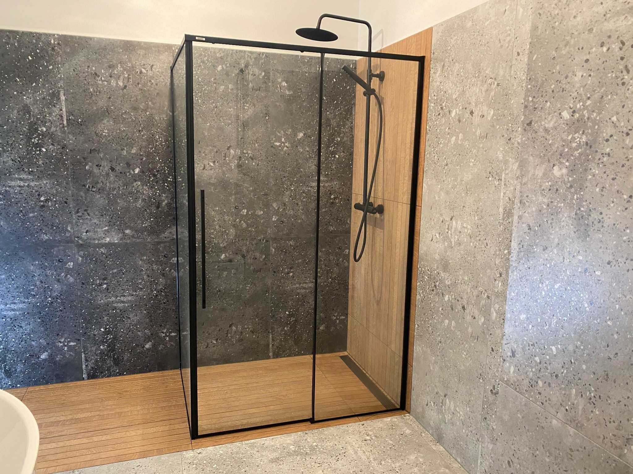 Nowoczesna kabina prysznicowa z czarnymi profilami, drewnianą podłogą i szarymi płytkami imitującymi lastryko na ścianach. Minimalistyczny design.