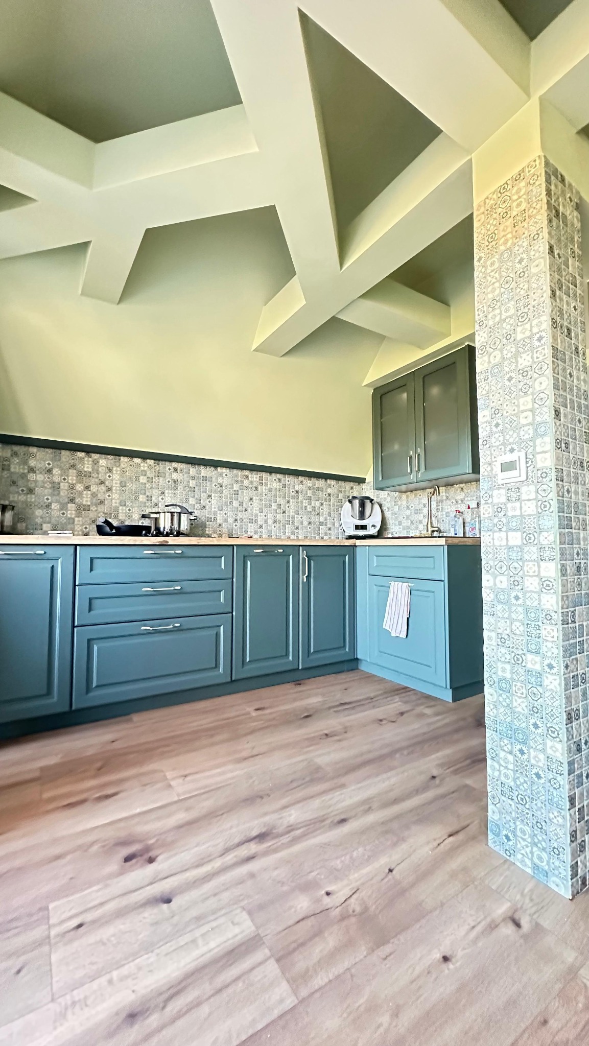 Turkusowa zabudowa kuchenna z drewnianym blatem i mozaiką nad blatem. Na podłodze panele. Sufit z dekoracyjnymi belkami. Kolumna wykończona ozdobnymi płytkami.