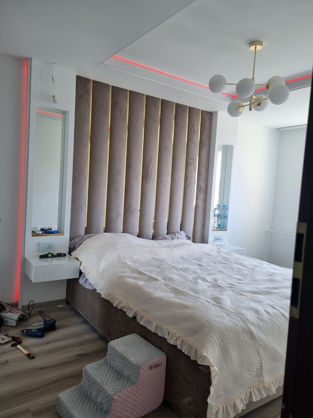 Sypialnia z łóżkiem i tapicerowanym wezgłowiem w pionowe pasy, oświetleniem LED przy suficie i lampą wiszącą. Na podłodze narzędzia i drabinka dla zwierząt.