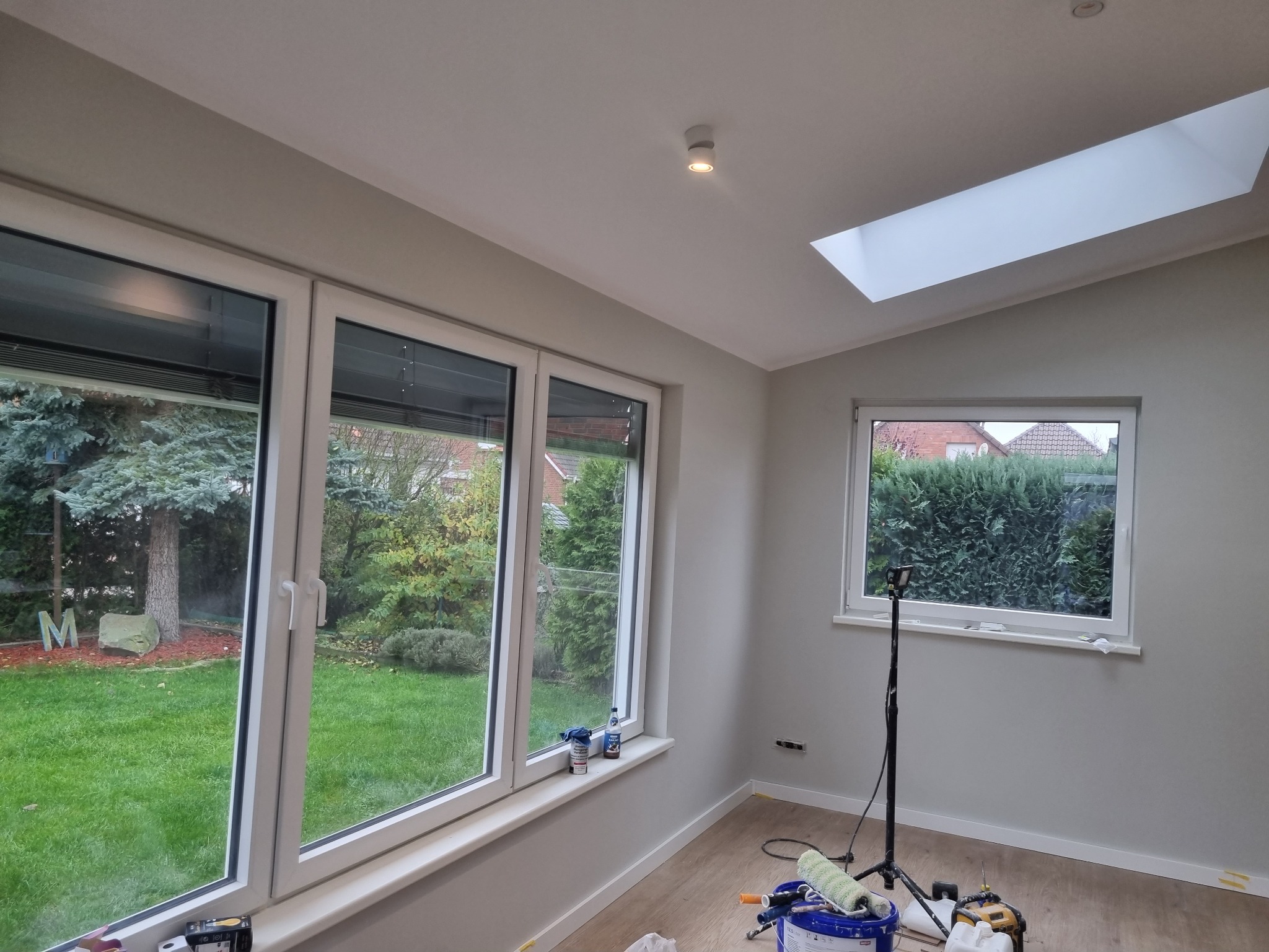 Pomieszczenie po malowaniu ścian w odcieniach szarości, z widokiem na ogród przez okna. Widoczne akcesoria malarskie i lampa oświetleniowa na statywie.