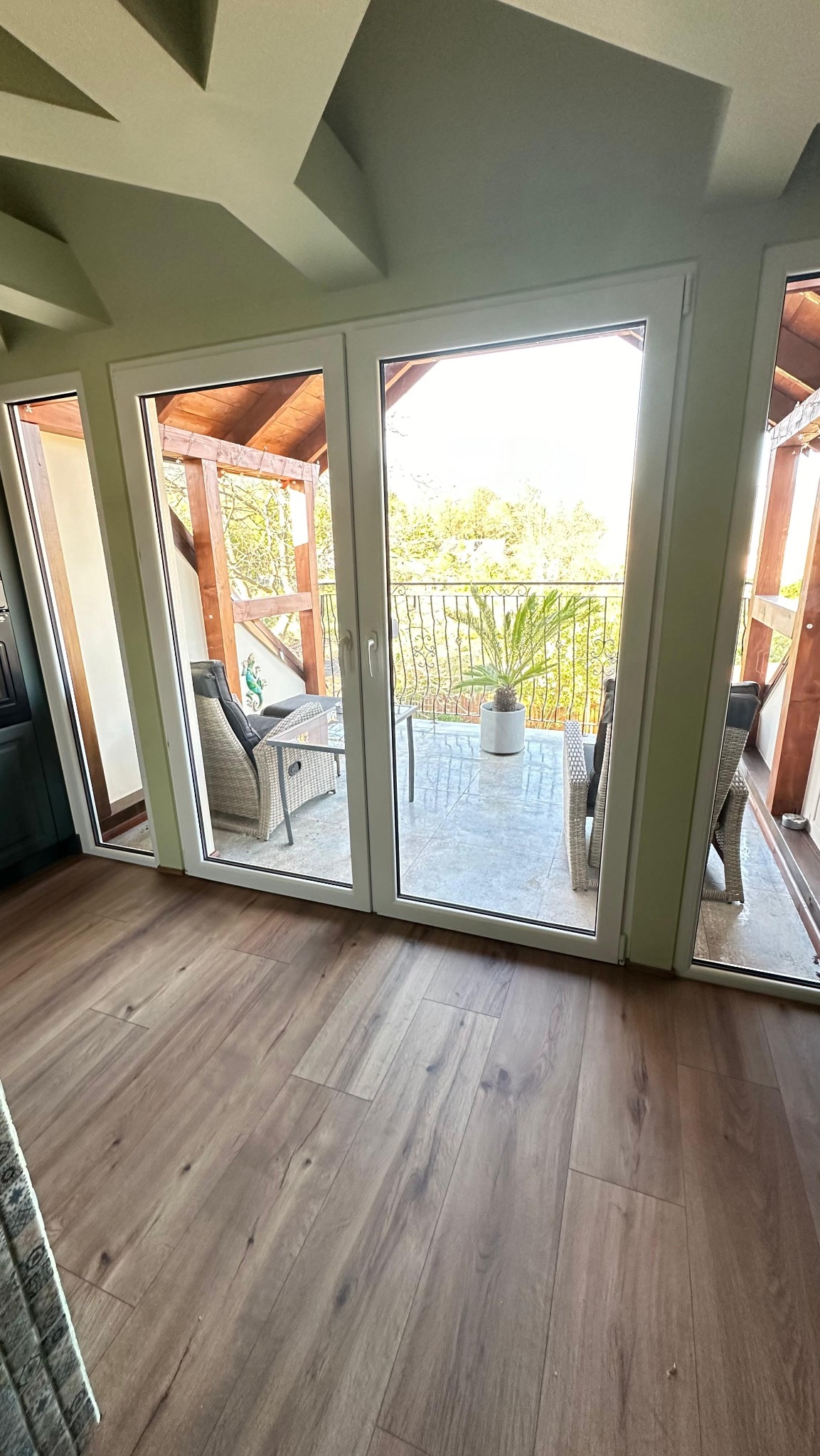 Jasne, drewniane panele podłogowe prowadzą do przeszklonych drzwi balkonowych. Widok na balkon z rattanowymi meblami i rośliną w donicy.