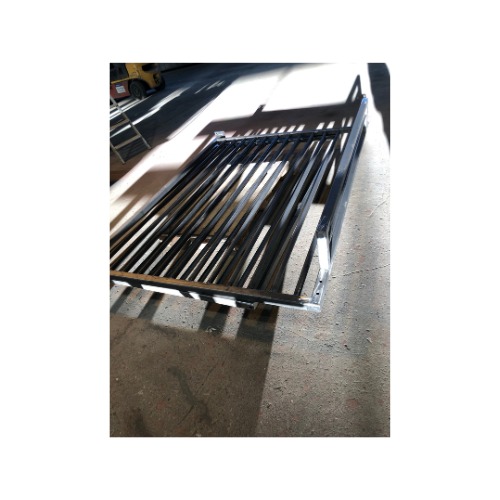 Metalowa konstrukcja z czarnymi prętami, spawana rama. Widoczne spawy i cień rzucany na podłoże. W tle wózek widłowy.