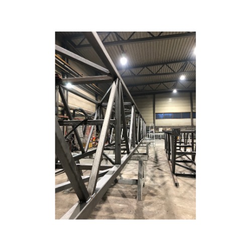 Metalowa konstrukcja kratowa, spawana, pomalowana na szaro, ustawiona na kozłach w hali produkcyjnej. Widoczne spawane łączenia i elementy konstrukcyjne.